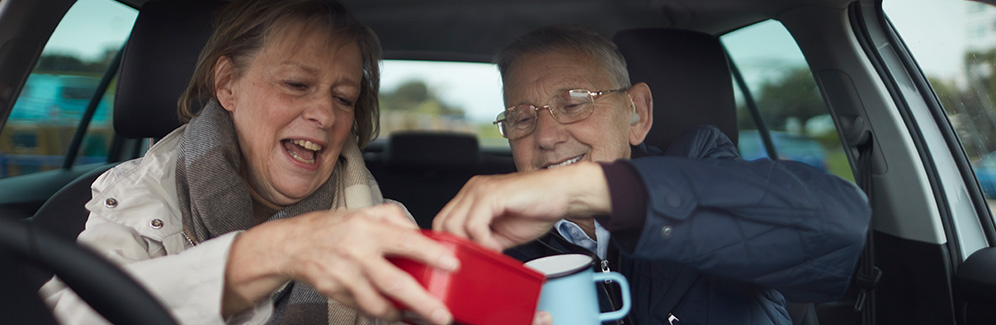 An older couple sat in the front of their car holding mugs and a red box. Accessibility features at South West